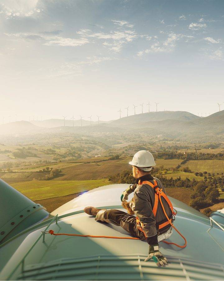 Technician in a hard hat and safety harness on top of a wind turbine above rolling farmland
